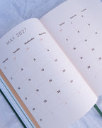 An open planner showing May 2027 on the left page and a weekly calendar from the 1st on the right page, photographed on a light textured fabric background.