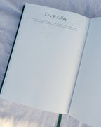 An open planner showing a blank page with the handwritten heading '2027 College' and a small faint line drawing, placed on a light blue textured fabric surface.
